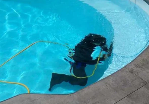 A person in black diving gear with a flashlight, inspecting the tile of a swimming pool underwater.