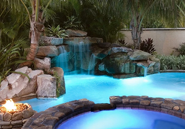 A swimming pool and spa with a rock waterfall and fire feature at dusk.