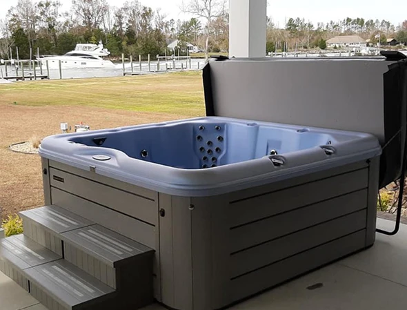 A large hot tub on a concrete patio with wooden steps, set in a backyard with a grassy hill and trees.