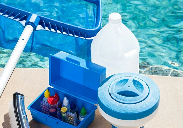 Pool cleaning supplies, including a skimmer net, chemicals, and test kit, by the edge of a swimming pool.