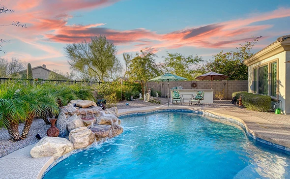 A large, modern swimming pool with a stone waterfall and steps, with palm trees and a house in the background, at sunset.