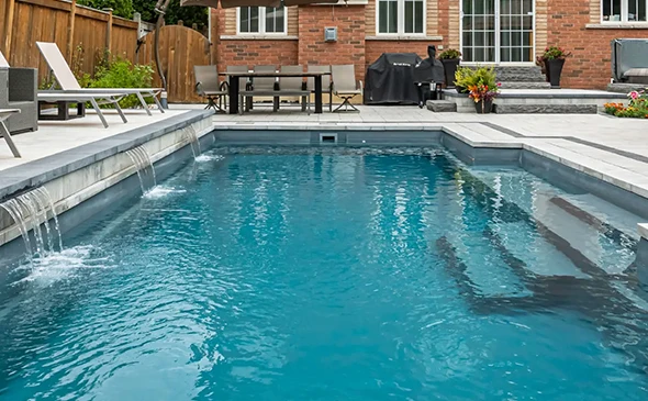A modern swimming pool with a built-in spa and stone coping, set in a backyard with a patio and landscaping.