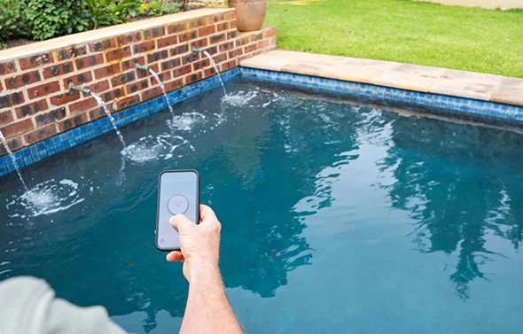 A person's hand holding a smartphone and controlling a pool system next to a pool with a water feature.
